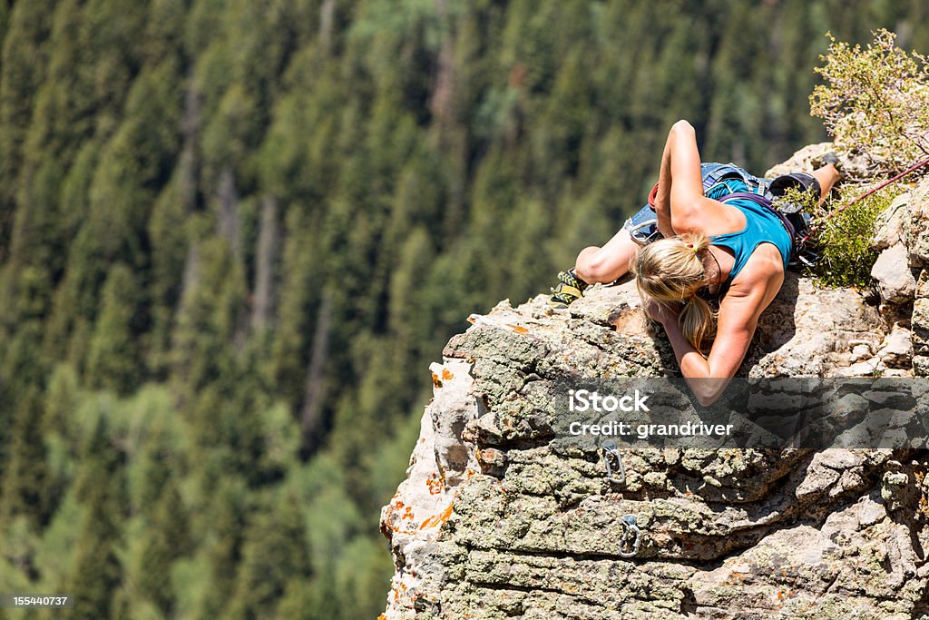 Female Mountain Climber on a Rock Face Caucasian female athletic blond rock climber on the sheer face of a granite cliff reaching up with her hands to gain more heights.  Subject is wearing all the appropriate climbing gear such as belts and carabiners to manage the safety rope.  Climber and cliff is positioned perfectly at the right third of the image revealing a pine tree forest in the distance creating ample copy space for titles and other design elements. Achievement Stock Photo