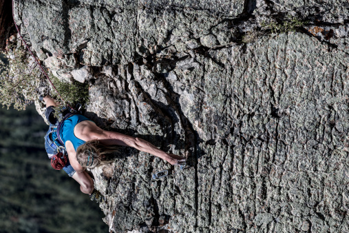 Caucasian female athletic blond rock climber on the sheer face of a granite cliff reaching up with her hands to gain more heights.  Subject is wearing all the appropriate climbing gear such as belts and carabiners to manage the safety rope.  Climber and cliff is positioned perfectly at the right third of the image revealing a pine tree forest in the distance creating ample copy space for titles and other design elements.
