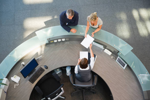 Business Couple At Reception Desk Elevated view of business couple standing at reception desk with receptionist handing over paper hotel reception hotel service technology stock pictures, royalty-free photos & images