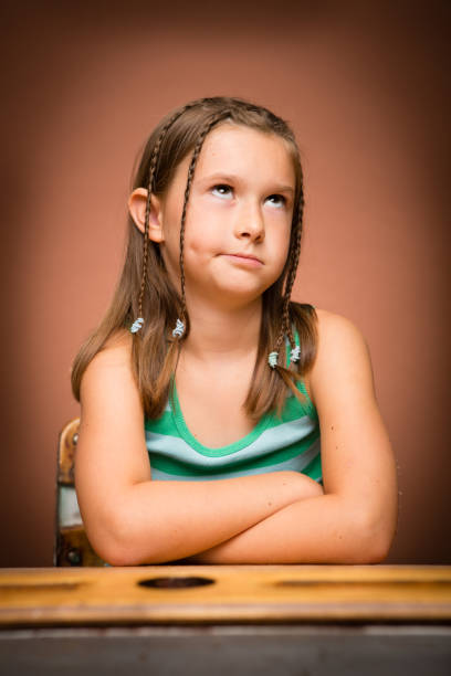 Color image of a little girl rolling up her eyes in disgust/impatience while sitting at a school desk. On a brown background.