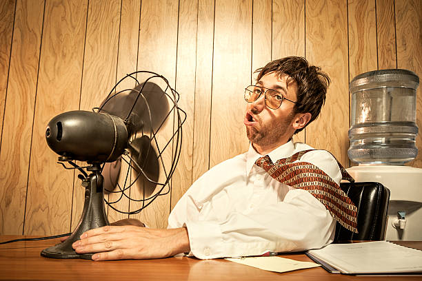 Business Man in Office With Fan A white collar man working in his office on a hot day sits in front of his fan, the wind blowing his tie, hair, and face as he tries to cool down.  Wood desk and wood paneling in the background, a water cooler visible in the background.  Horizontal with copy space. electric fan photos stock pictures, royalty-free photos & images