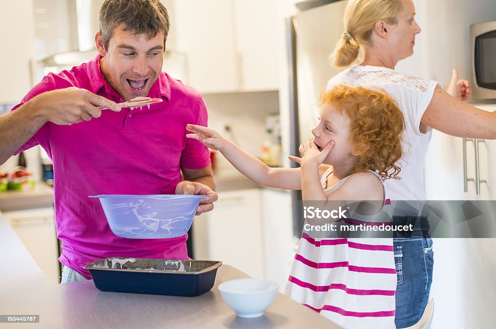 Real family of three making cake in the kitchen. Dad, mom and their little girl are making a banana-chocolate cake in the kitchen. Daughter and dad are fighting about who is going to lick the spoon…Natural light and shallow DOF, focus on the little girl. 30-34 Years Stock Photo