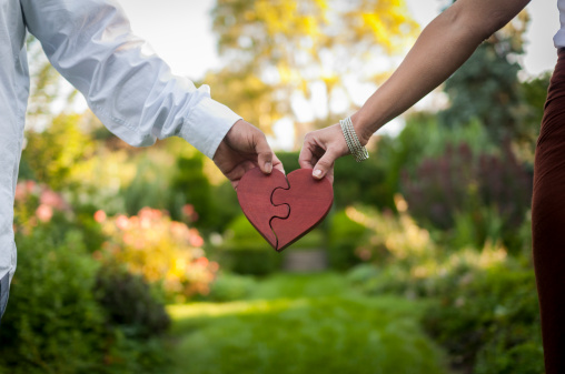 Man and woman's hands holding onto a two piece heart shaped puzzle. Out of focus garden in background.