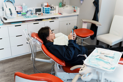 Female patient of Asian ethnicity watches her phone as she lays in a dental chair, waiting to start her examination.