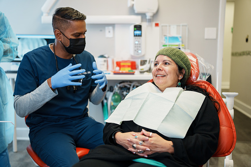 White, female patient sits in a dental chair smiling, as she builds trust and consults with a male dentist of Indian descent before check up.