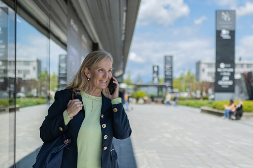 A business woman, casually dressed, walks through the streets of the city, in a good mood, smiling, talking on the phone and enjoying a sunny day. A woman walks by the glass store and business windows.Copy space, portrait.