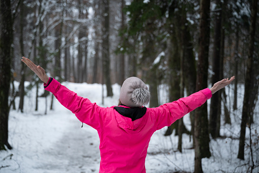 View from behind of a woman in pink sweater standing in calm snow covered winter forest with her arms spread. Enjoying and celebrating life.