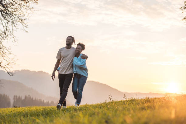 Carefree Love: A Young Couple enjoying a Spring Day Young black couple is hugging while smiling and walking in a green hill during the sunset. They are dressed in casual outfits and taking in the beauty of nature. black couple sunset stock pictures, royalty-free photos & images