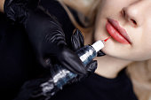 Cosmetologist applying red permanent make up tattoo on young woman lips. Permanent lips tattoo procedure concept.