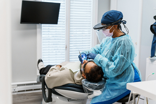 Multiracial, female patient sits, performing a tooth filling procedure on a male patient of Indian descent.