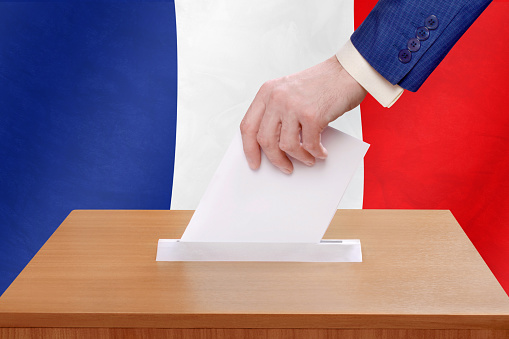 Election in France. The hand of man putting his vote in the ballot box. French flags on background.