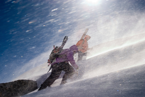 Two back country riders breaking a trail up, on a steep mountain slope to the a summit. They are moving slowly fighting with the wind gusts.