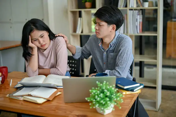 A caring and kind young Asian man is comforting and showing support to his upset and stressed-out girl friend while studying and preparing for the exam in a campus library together.