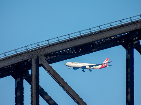 A Qantas Airbus A330-303 plane, registration VH-QPD, approaches the main runway of Sydney Kingsford-Smith Airport as flight QF26 from Tokyo, passing the Sydney Harbour Bridge in the foreground.  This image was taken from Mrs Macquarie's Chair on a sunny, cold morning on 19 July 2023.