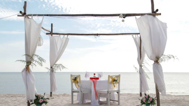 istock Romantic dinner table set and chair preparation with white theme decoration, white sand, clear cloudy sky, and tranquil, peaceful, beautiful beach view at the seashore. Good place for wedding proposal 1554931826