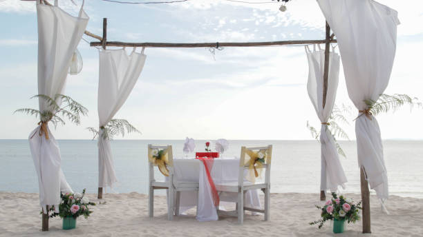 istock Romantic dinner table set and chair preparation with white theme decoration, white sand, clear cloudy sky, and tranquil, peaceful, beautiful beach view at the seashore. Good place for wedding proposal 1554931839