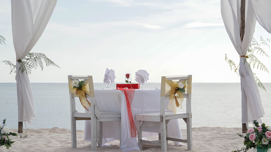Romantic dinner table set and chair preparation with white theme decoration, white sand, clear cloudy sky, and tranquil, peaceful, beautiful beach view at the seashore. Good place for wedding proposal