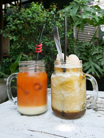 Iced lychee tea and Iced Thai tea served on a white table with a background of tropical plants and old rustic teak wood.