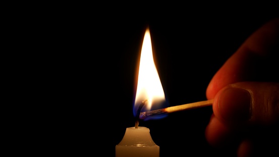 Close-up of a man's hand lighting a wax candle with a match against a black background. Bright flame, glowing fire. Concept of remembrance, ritual, remembrance, celebration or religious ceremony