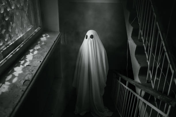 istock Ghost covered with a white sheet walks through an abandoned building and looks out the window. Black and white photo 1555121322