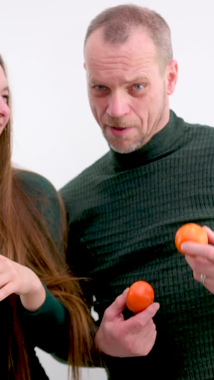 young girl put Tangerines to eyes and pulled out tongue stuck out and she twists and makes faces in the background a man on a white background a green sweater long hair joke fooling around grimacing