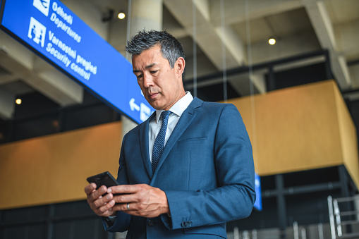 Asian businessman at departures terminal. Standing in a lobby using a smartphone.