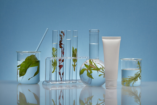 An empty white plastic tube decorated with laboratory equipment and fresh seaweed leaves on a backlit blue background .Mockup packaging, cosmetics design. Advertising photo