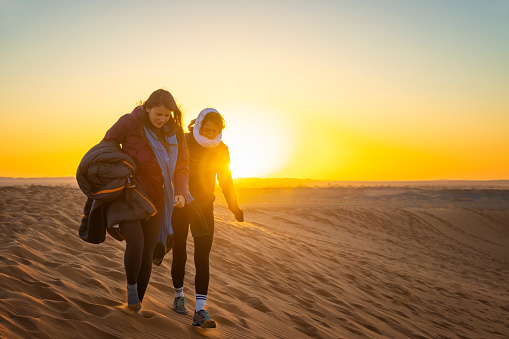 Woman tourists walking at sunrise on sand dunes in the desert, Merzouga, Erg Chebbi sand dunes region, Sahara, Morocco.