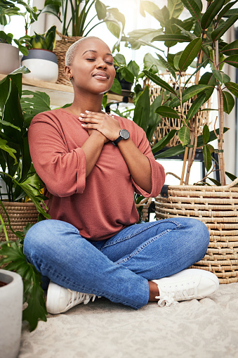 Calm, peace and young woman by plants for breathing exercise in meditation in a greenery nursery. Breathe, gratitude and African female person with a relaxing zen mindset by indoor greenhouse garden.