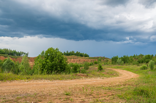 Dirt sandy road among the trees under a stormy sky on a summer day. Beautiful landscape of Russian nature.