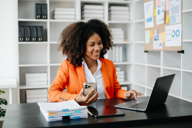 istock Young African woman typing on tablet and laptop while sitting at the working black table office 1555311032