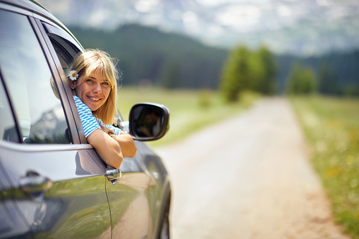 Beautiful blonde woman smiling with flower in her hair. Rear view of car. Transport, roadtrip, nature concept.
