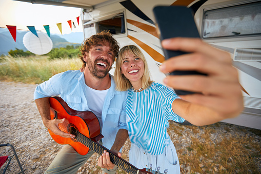 A young couple is taking a selfie while playing guitar at the campsite on a beautiful day in the nature. Relationship, vacation, nature, camping