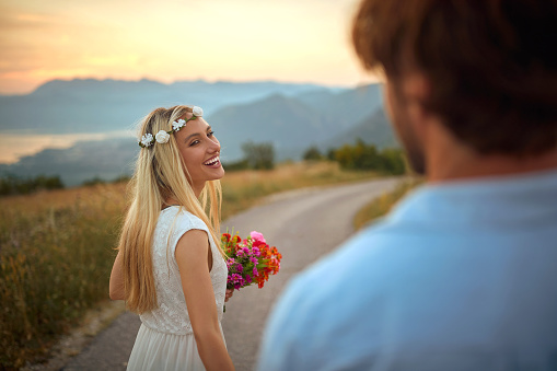 portrait of beautiful blonde turned around in motion with red bouquet in her hands and wreath of flowers on her head, smiling, laughing, looking at her fiancé