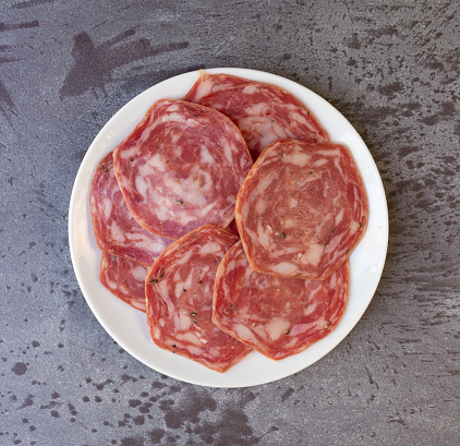 Top view of a group of slices of sweet sopressata on a white plate atop a gray mottled tabletop illuminated with natural lighting.