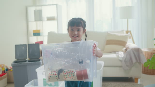 Asian girl kid holding a box full of plastic bottles to recycle happy and smiling looking at camera in living room at home. Family happy moment.