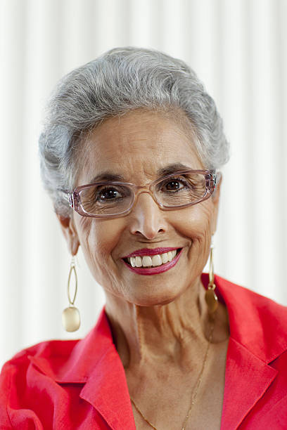 Portrait of senior Caucasian woman standing at podium addressing audience, short gray hair, serious expression, flags of multiple countries visible in background, formal setting