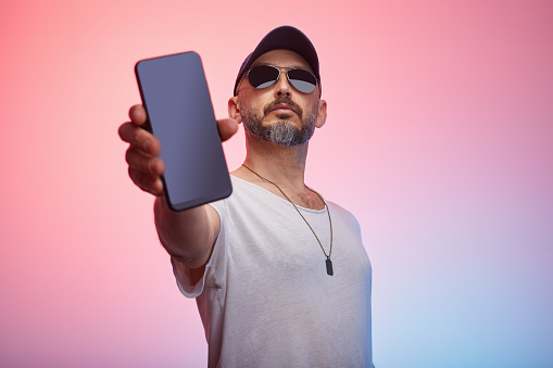 Studio portrait of a man wearing casual clothing, t-shirt and a cap shows blank screen of smart phone in front of colorful background