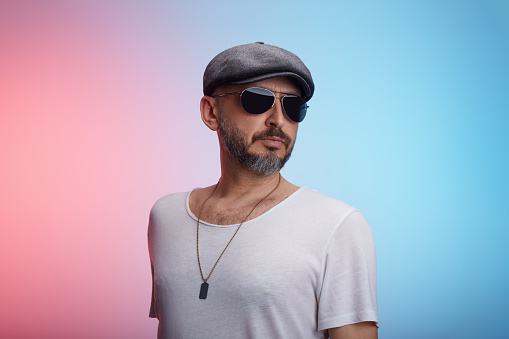 Studio portrait of a middle-aged confident man wearing a t-shirt, cap and sunglasses posing against a colorful background