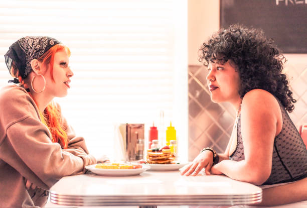 Two non-binary friends chatting as they sit face to face at a cafe.