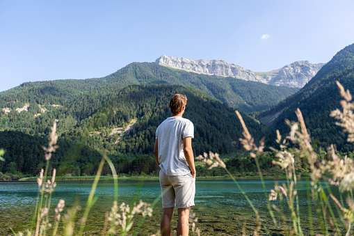 Boy up to his feet in a lake, with his hands in his pockets and looking at the mountains.