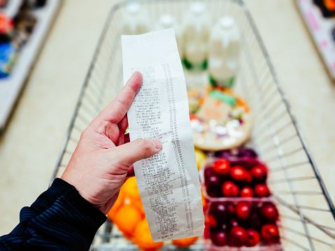 Holding and checking the bill after buying groceries in the supermarket. Close up of the receipt with the groceries defocused in the trolley below.