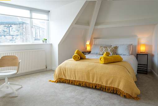 A comfortable double bed is set in an attic room. On the bed are towels and a bright orange over blanket. On each side of the bed, on a bedside table is an orange lamp in a tubular form. The carpet is a light grey colour. A large window and a desk chair completes the scene.