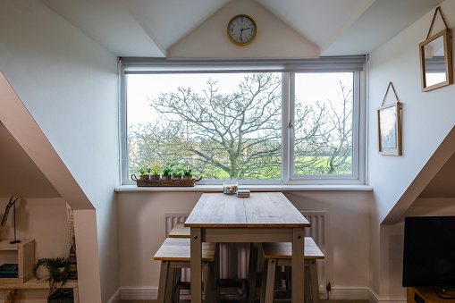 A view of a high table and four stools set against a window in an attic flat overlooking a budding tree with a green space beyond.