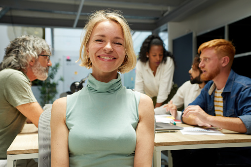 Portrait of an office cheerful Nordic mature woman and looking at camera. Empowered female leader. In the background there is a group of employees kipping at work. It is an indoor multiracial scene.