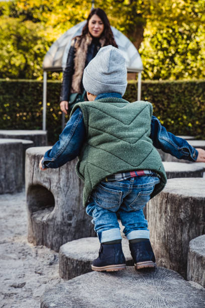 A small kid is jumping with energy from a tree stump while his proud mom is watching. Family day at the playground. Mother and son in a park as the child is hopping energetically among trunk stumps