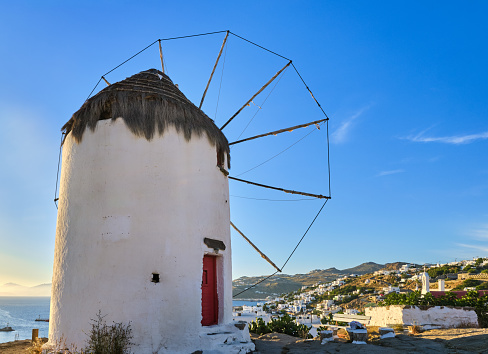Famous tourist attraction of Mykonos, Cyclades, Greece. Traditional whitewashed windmill on hill top overlooking town of Chora by waterfront. Summer, sundown, blue sky. Travel destination, iconic view.