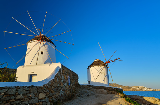 Famous tourist attraction of Mykonos, Cyclades, Greece. Two traditional whitewashed windmills by waterfront and Chora town. Summer, morning, clear blue sky, travel destination, iconic view. Frontal shot.