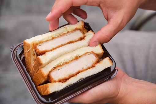 Unrecognizable person eating katsu sando, Japanese sandwich with pork cutlet, cabbage and tonkatsu sauce, at street.