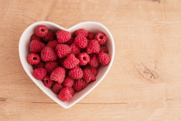 istock Fresh raspberries lie in a white heart-shaped salad bowl. There is a table surface made of light wood. Flatley. Background. 1555680927